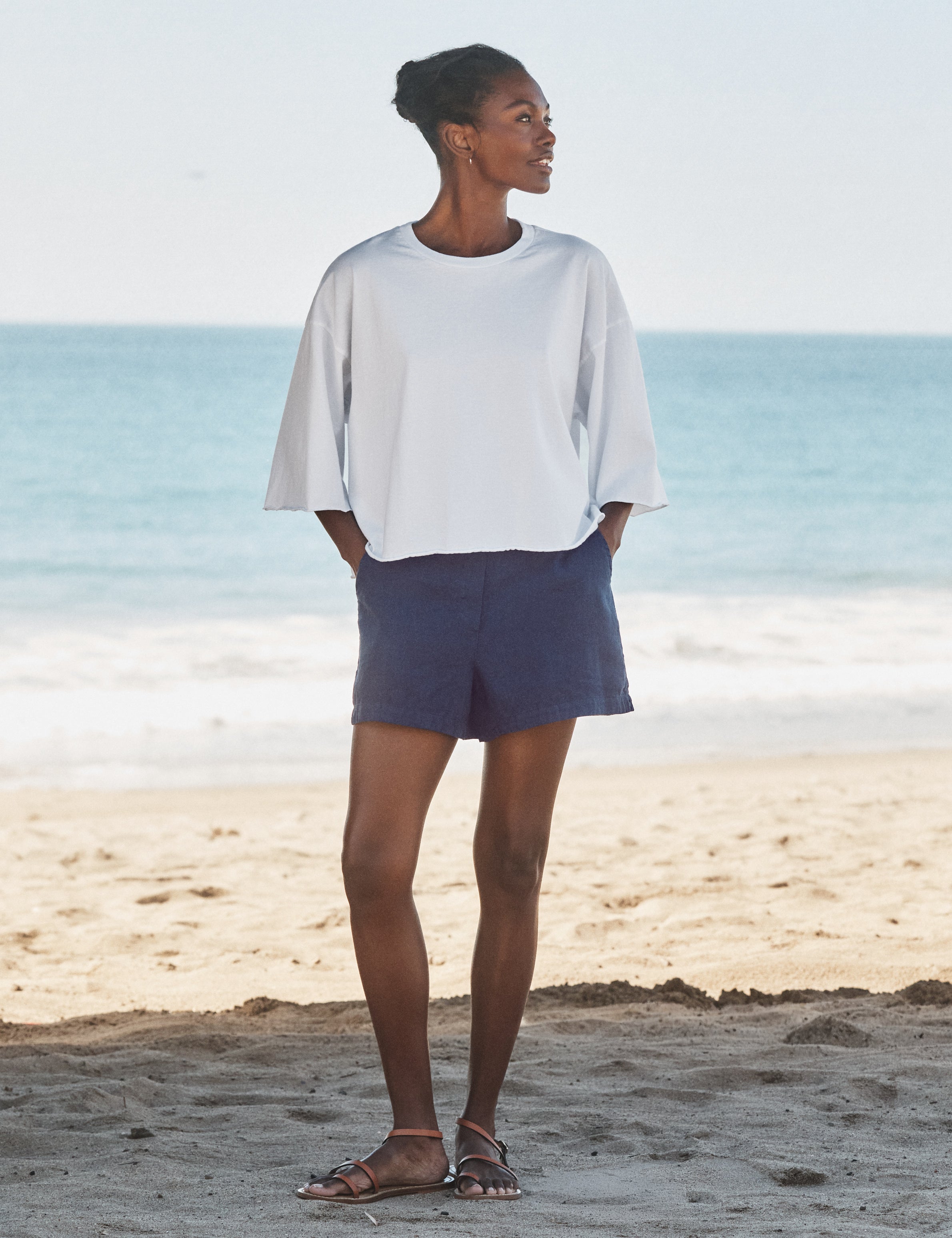 Woman standing on a beach wearing a white top and navy shorts., view 4