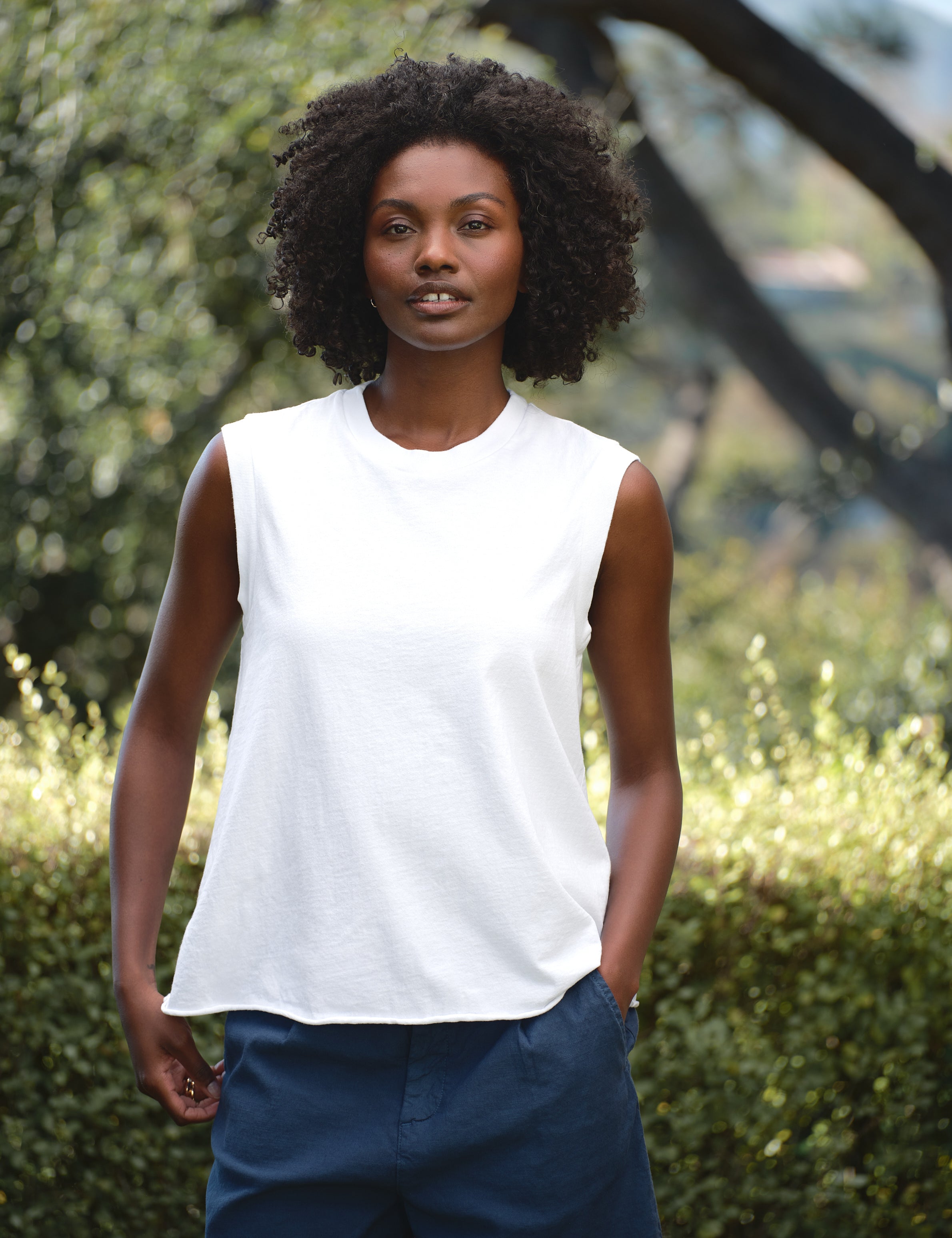 Woman wearing a white sleeveless top and blue pants standing outdoors with greenery in the background, view 3
