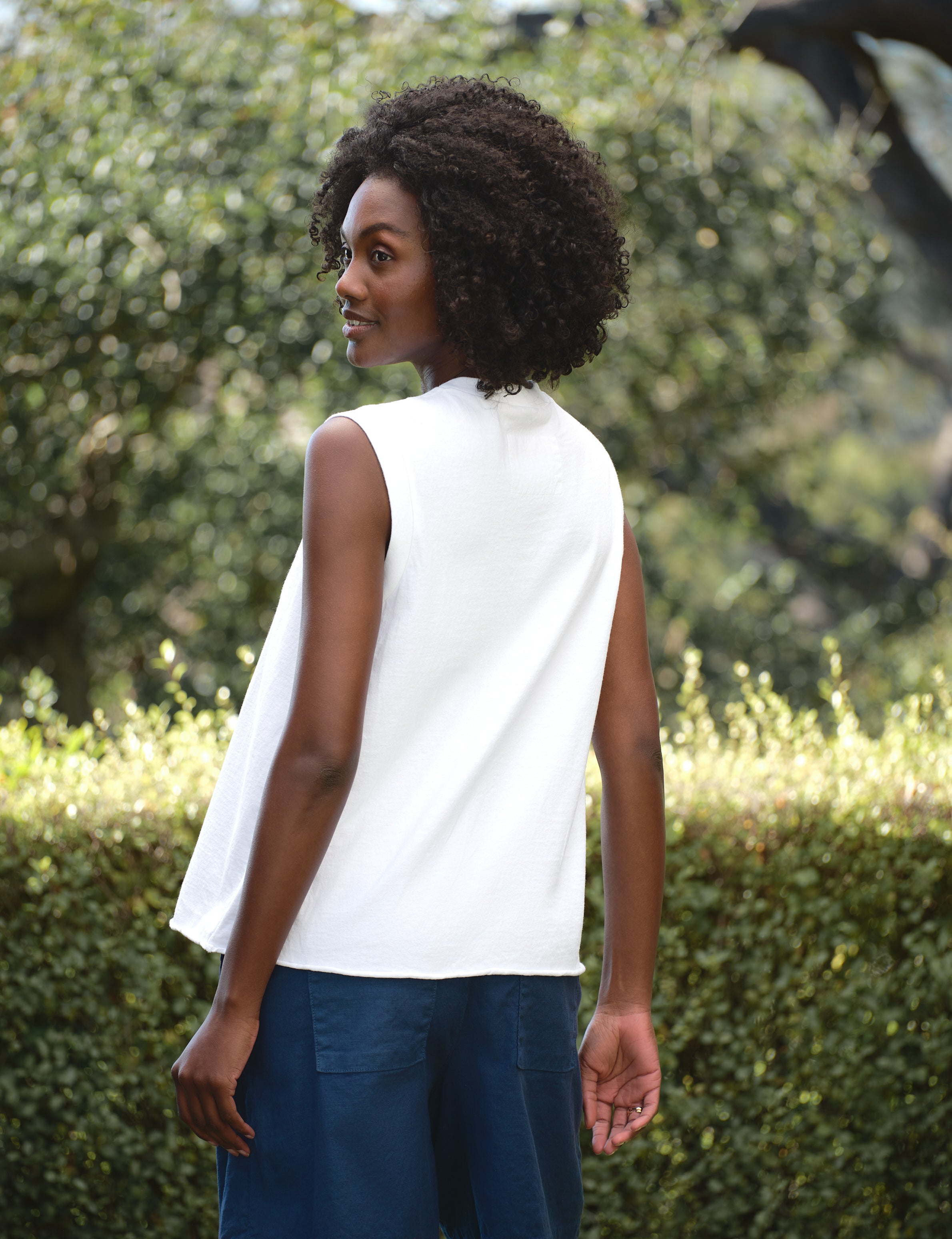Woman wearing a white sleeveless top and blue shorts standing outdoors with greenery in the background, view 4