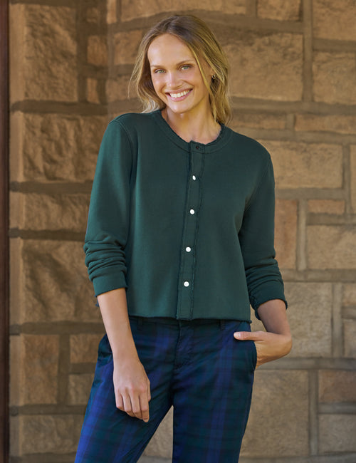 A woman wearing a dark green long-sleeve button-front top and blue plaid pants, standing against a tan stone wall., front view