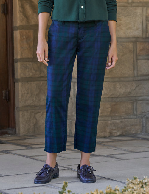 A person wearing cropped plaid trousers in dark blue and green tartan, paired with a dark green top and dark lace-up shoes, standing outdoors against a stone wall., front view