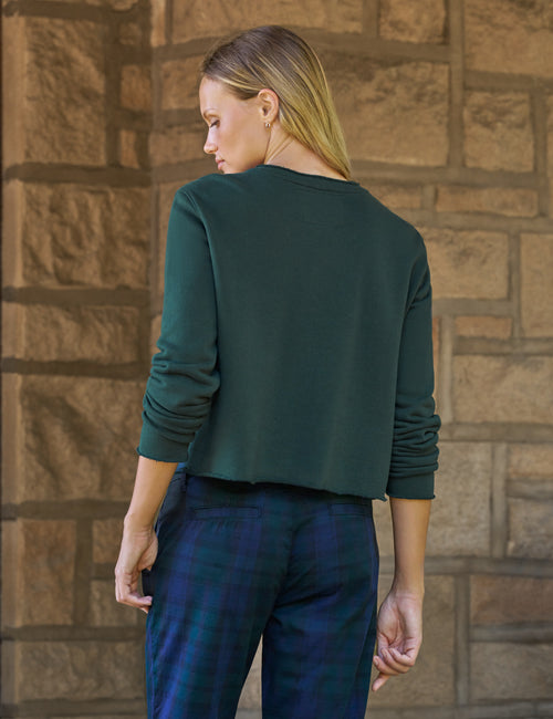 A woman shown from the back wearing a dark green long-sleeve cropped top and blue plaid pants, standing against a brown stone wall., alternate view