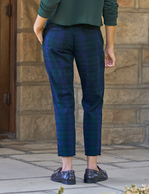 A person shown from the back wearing dark blue and green plaid cropped trousers, a dark green top, and dark brown leather shoes, standing outdoors against a stone wall., alternate view