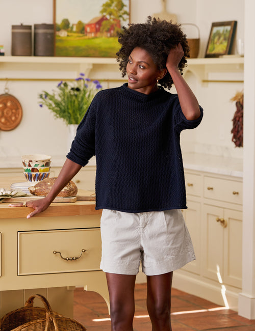 Woman standing in a kitchen wearing a navy sweater and tan shorts., view 5