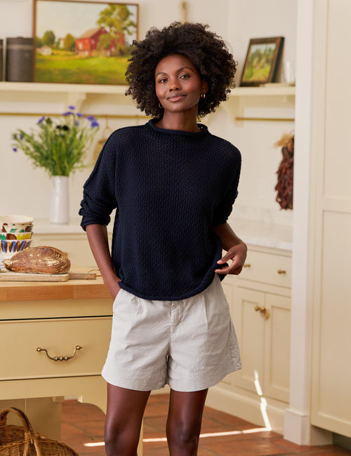 Woman standing in a kitchen wearing a navy sweater and light shorts., view 4