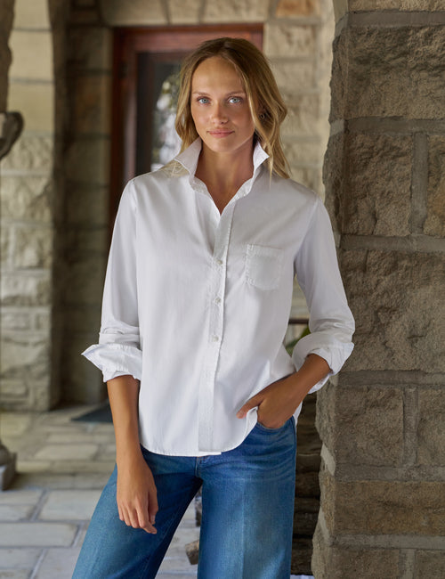 A woman wearing a white long-sleeve button-down shirt with rolled cuffs and blue jeans, standing in a stone corridor., front view
