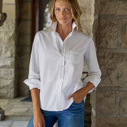 A woman wearing a white long-sleeve button-down shirt with rolled cuffs and blue jeans, standing in a stone corridor.