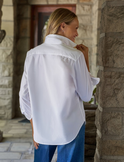 A woman wearing a white long-sleeve button-up shirt and blue jeans, standing in front of a stone pillar and doorway., alternate view
