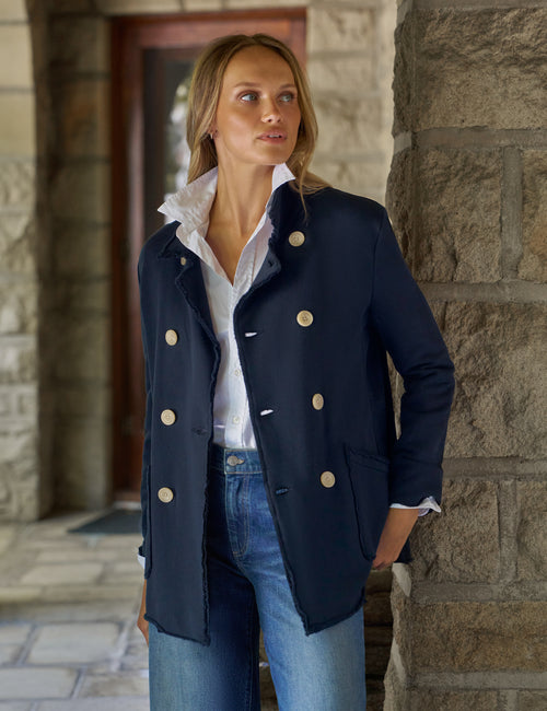 A woman wearing a navy double-breasted blazer with tan buttons, a white collared shirt, and blue jeans, standing in front of a stone column., front view