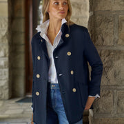 A woman wearing a navy double-breasted blazer with tan buttons, a white collared shirt, and blue jeans, standing in front of a stone column.