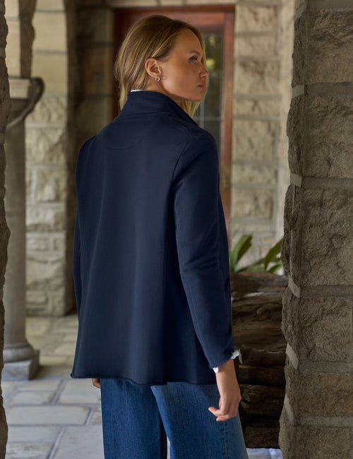 A woman wearing a navy blue blazer and blue jeans, standing on a stone porch and looking to the side., alternate view