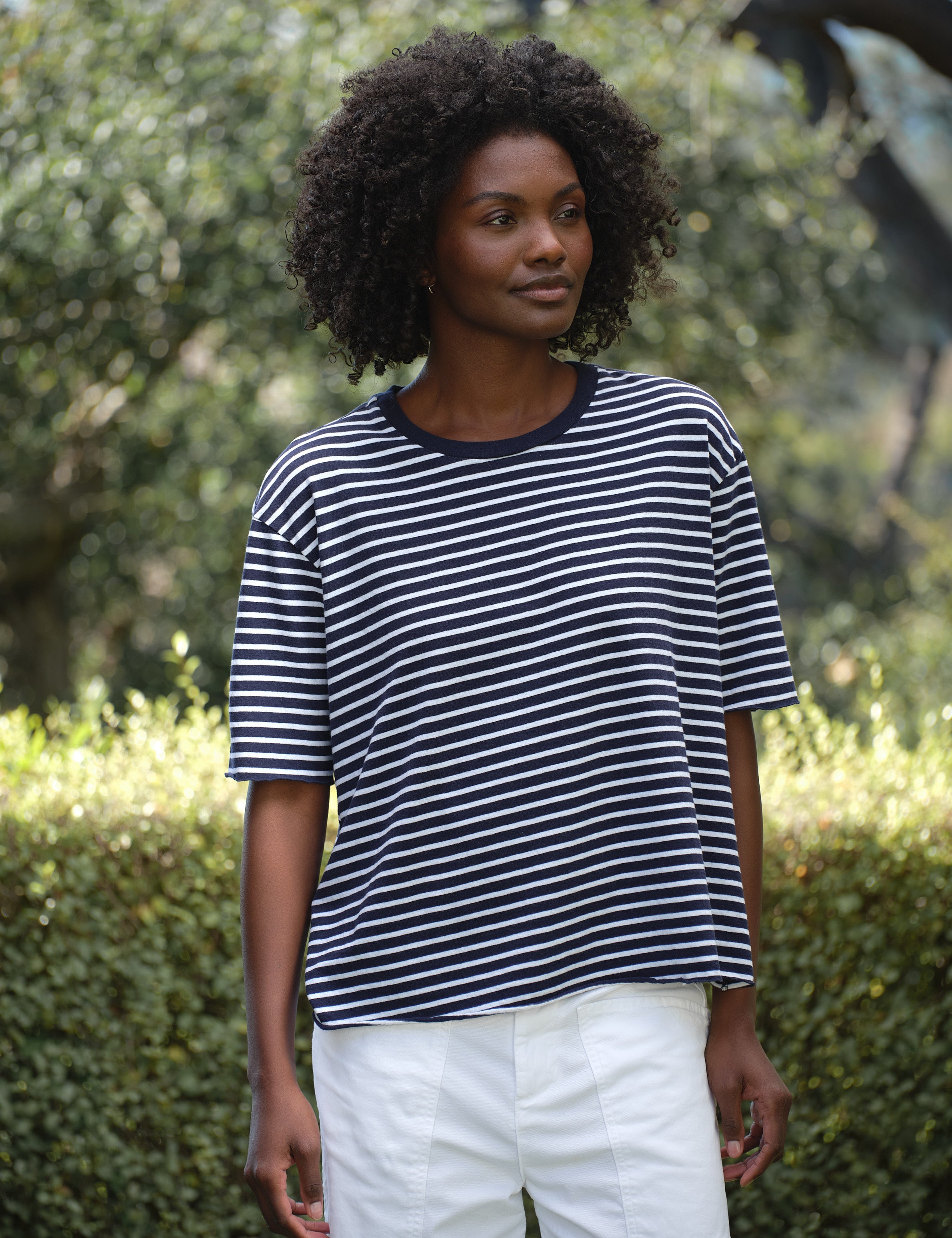 Woman wearing a striped shirt and white pants standing outdoors with greenery in the background, front view