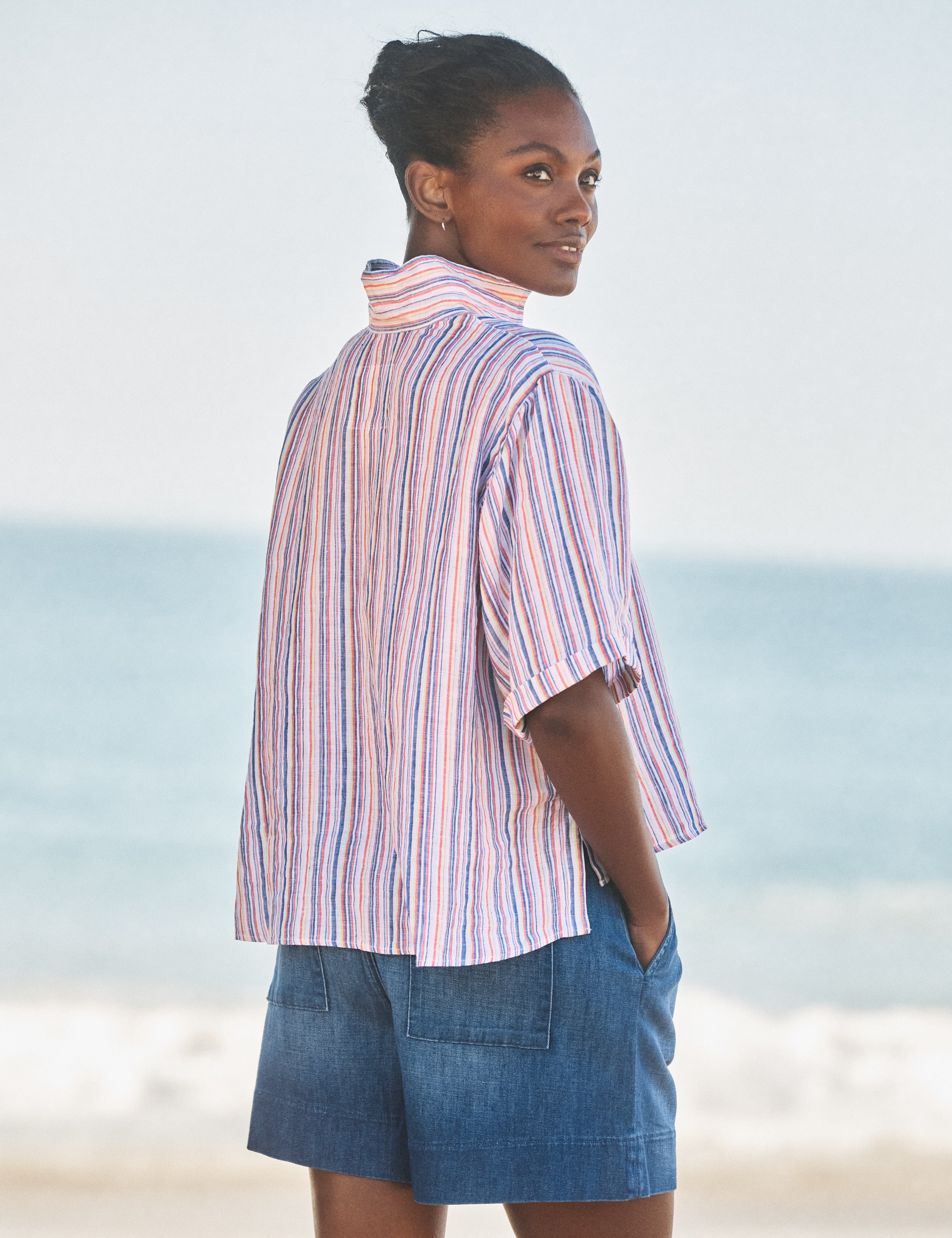 Woman wearing a striped shirt and denim shorts standing on a beach., alternate view