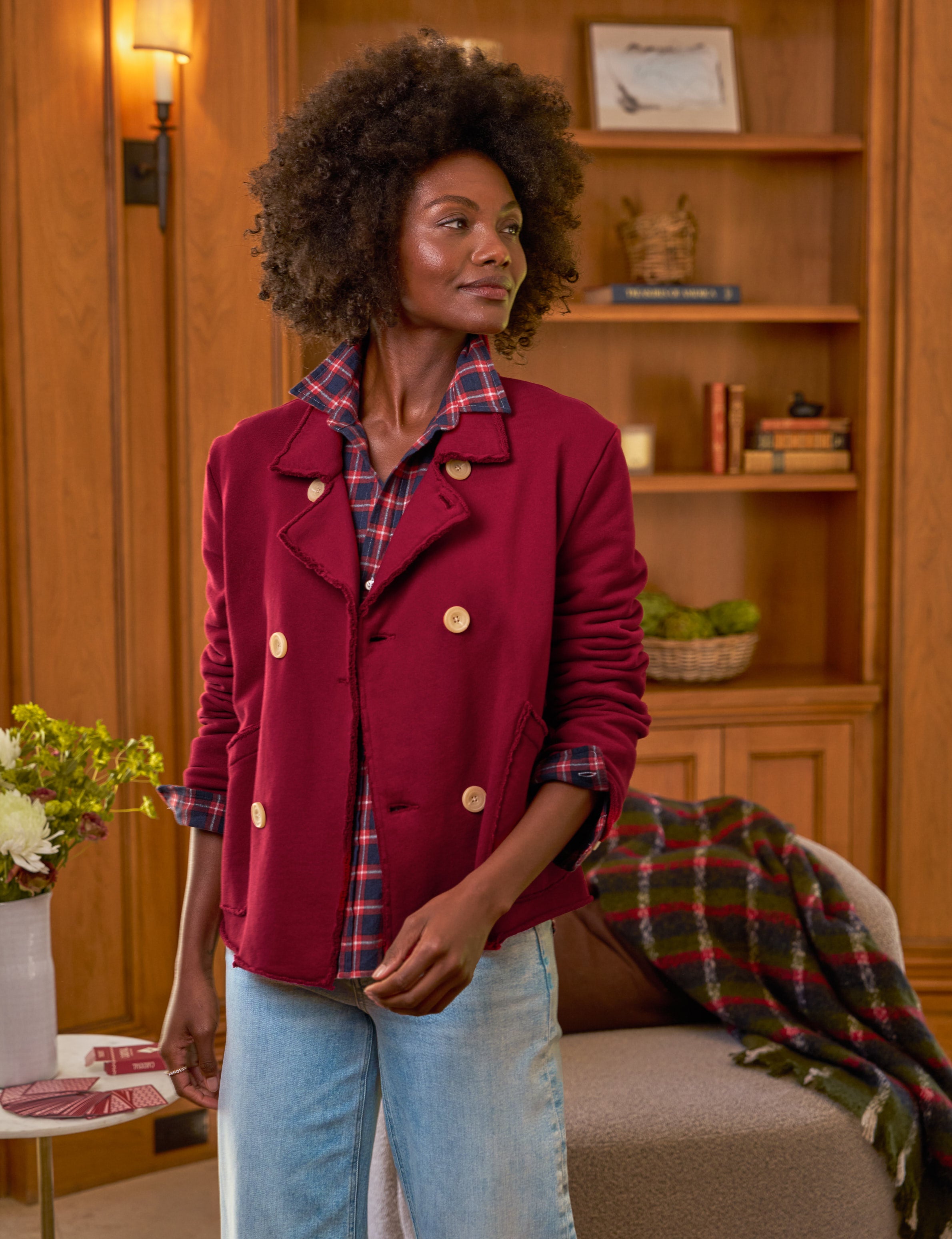 Woman wearing a red jacket with a plaid shirt and blue jeans in a cozy room.