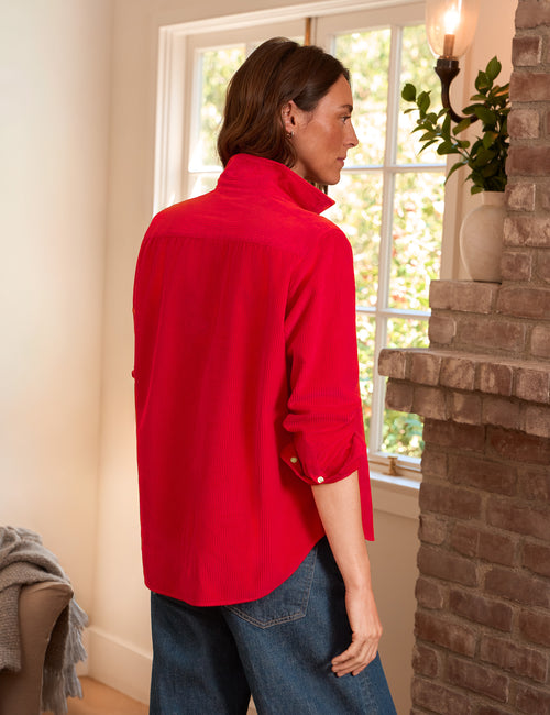 A woman wearing a bright red long-sleeve blouse with rolled cuffs and blue jeans, standing in a warmly lit room next to a brick fireplace and window., alternate view