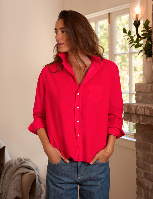 A woman wearing a bright red button-up shirt with rolled sleeves and blue jeans, standing indoors near a window and brick fireplace., front view