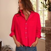 A woman wearing a bright red button-up shirt with rolled sleeves and blue jeans, standing indoors near a window and brick fireplace.