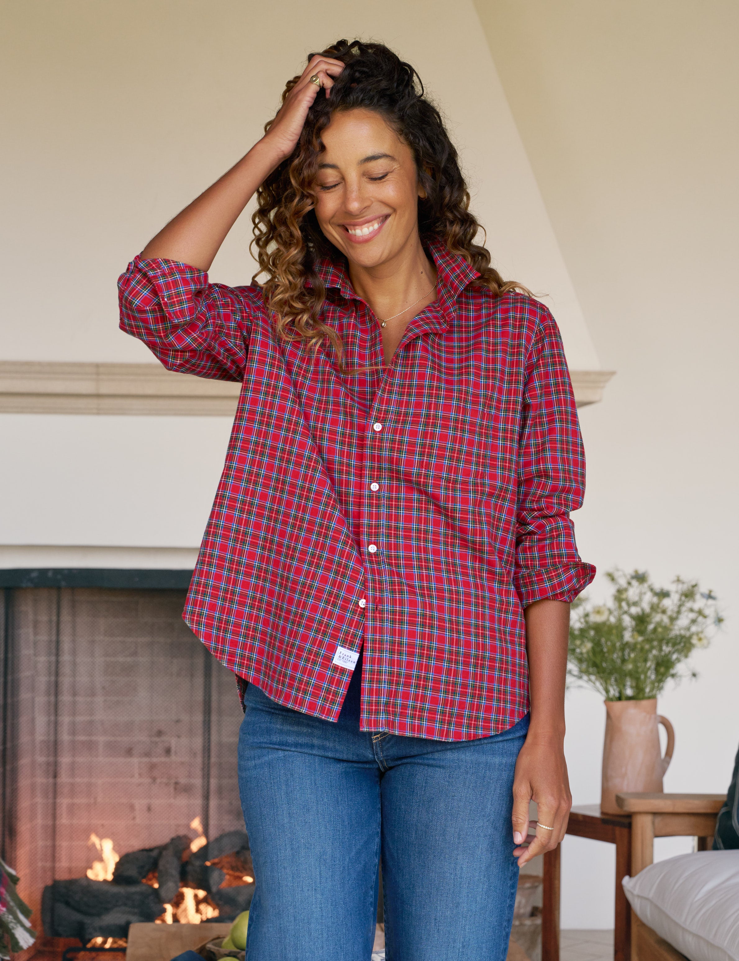 Woman wearing a red plaid shirt and blue jeans standing in a cozy living room with a fireplace.