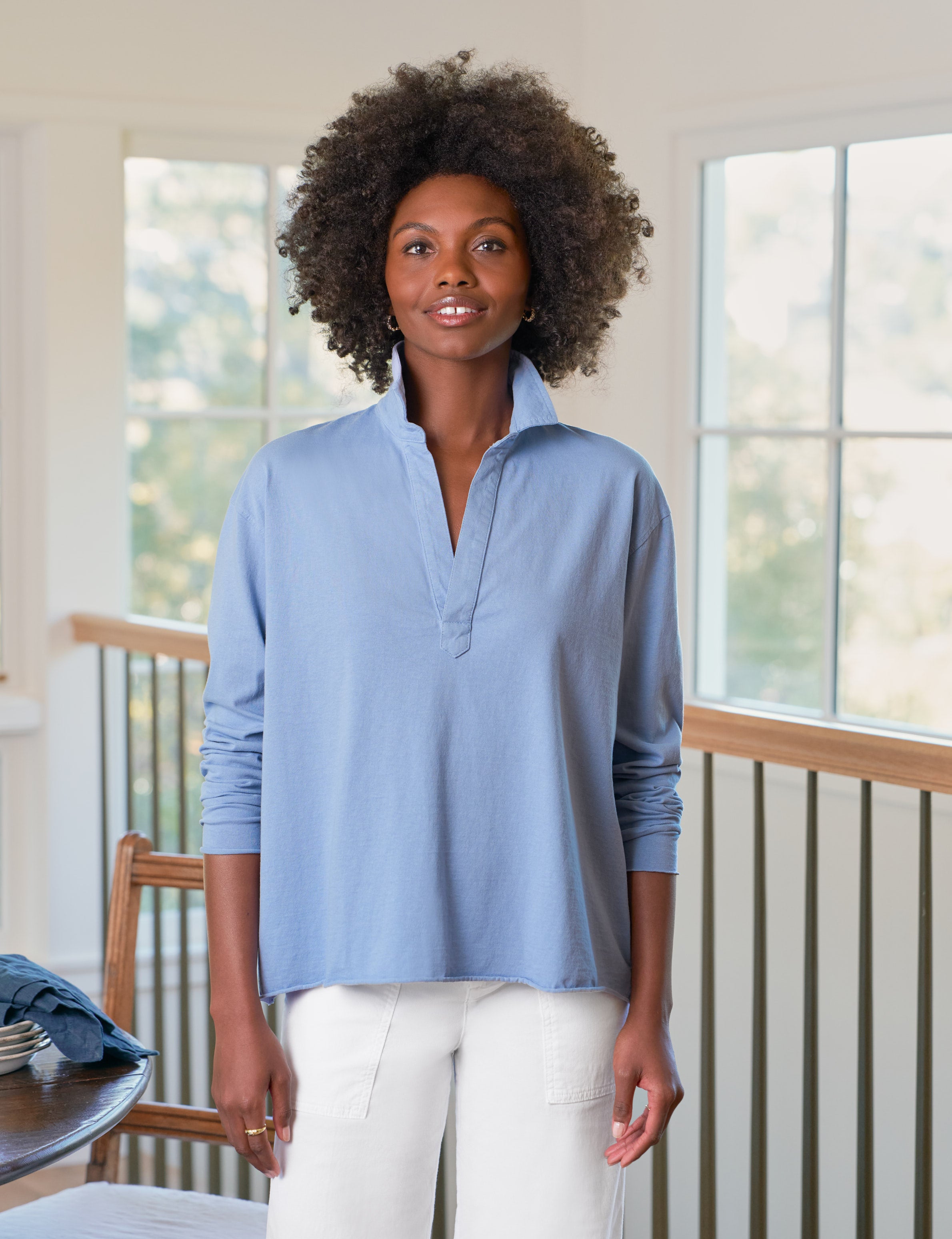 Woman wearing a light blue shirt and white pants standing in a room with large windows.