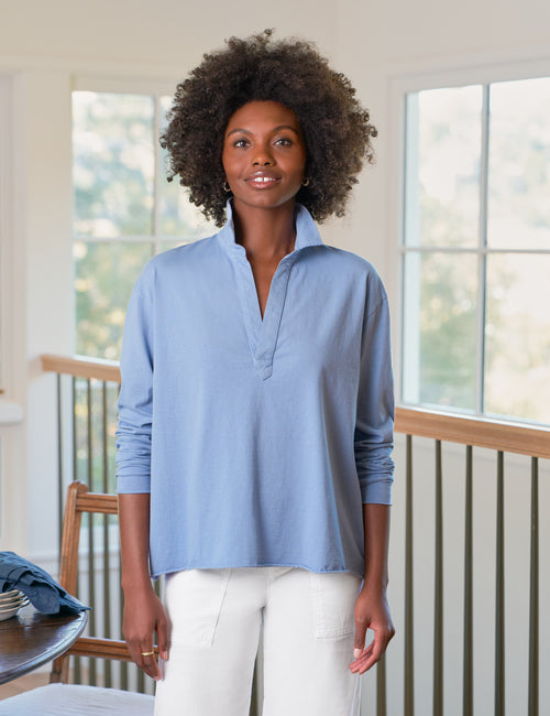 Woman wearing a light blue shirt and white pants standing in a room with large windows., front view
