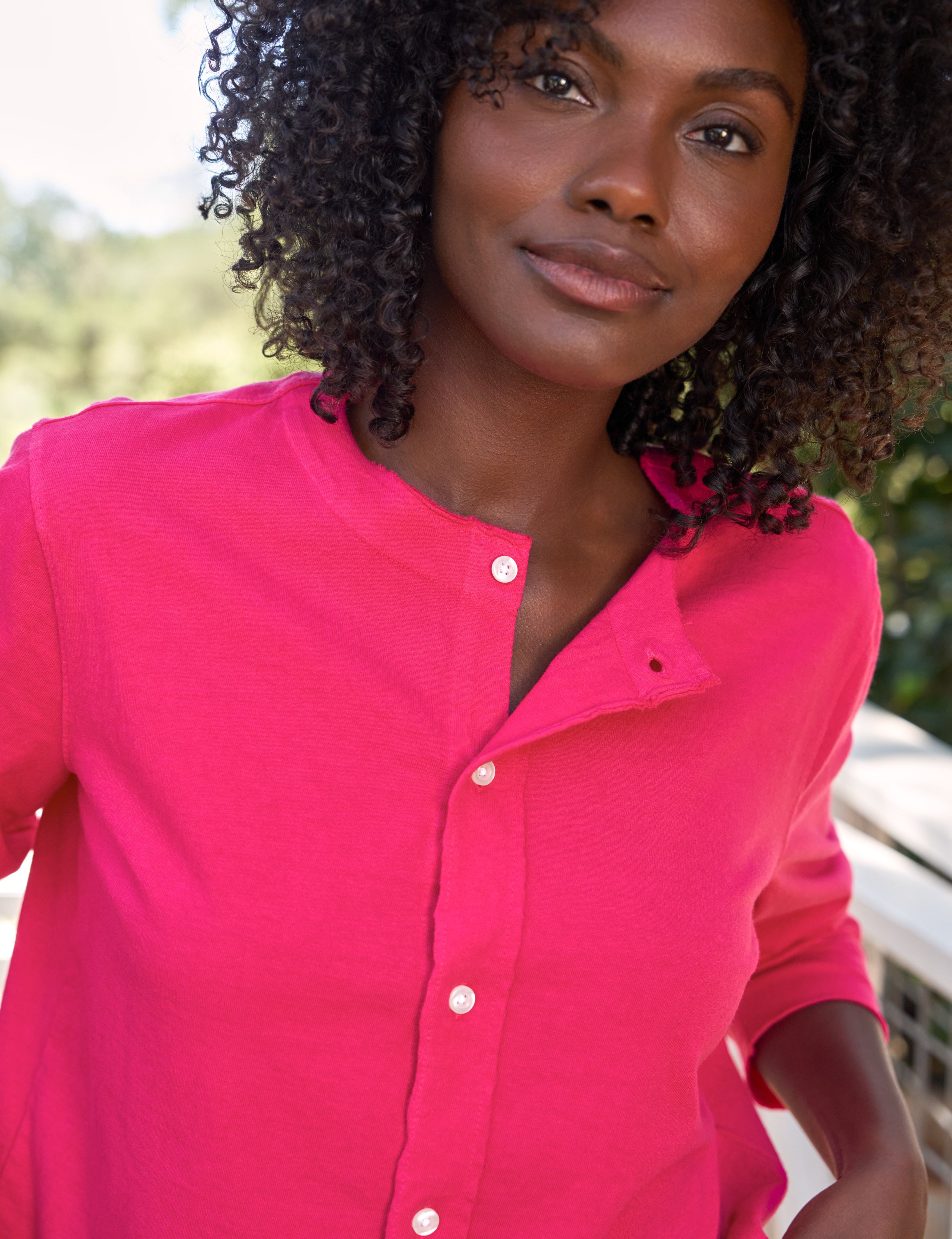 Woman wearing a bright pink shirt with a blurred outdoor background, view 3