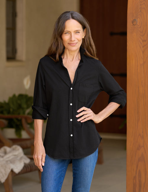 A woman wearing a black button-down shirt with rolled-up sleeves and blue jeans, standing in a doorway., front view