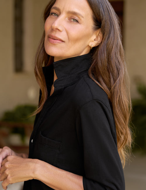 A woman with long brown hair wearing a black high-neck button-up shirt, shown in a close-up profile view against a softly blurred outdoor background., view 3