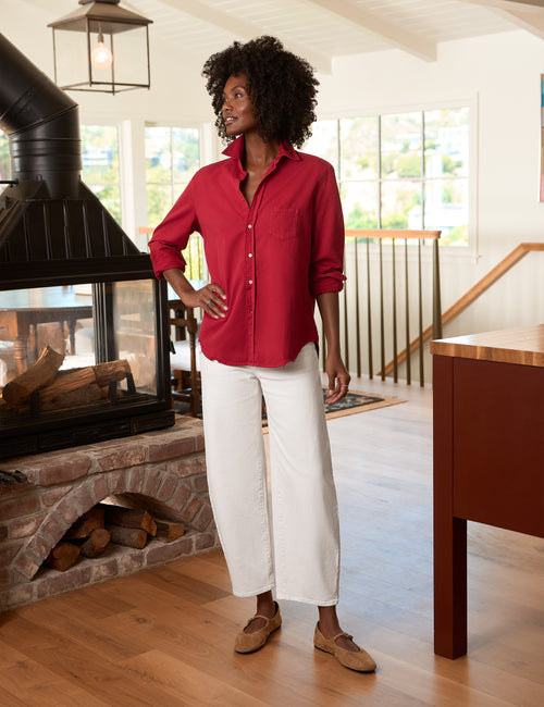 A woman stands indoors wearing a bright red button-up shirt, white cropped pants, and tan loafers. The setting is a modern home interior with a fireplace, wood floors, and large windows., view 4