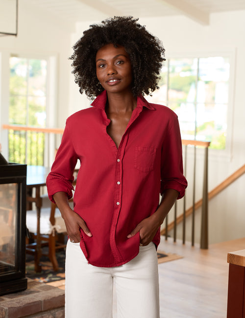A woman with curly black hair wearing a red button-up shirt and white pants, standing in a bright, modern home interior with a staircase and windows in the background., alternate view