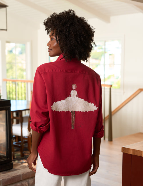 A woman with curly black hair is shown from the back wearing a red button-up shirt with a white tutu and ballerina design on the back, paired with white pants in a bright indoor setting., front view
