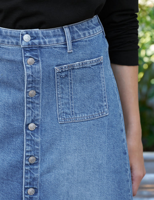Close-up of a light blue denim skirt with a button-front closure and a front patch pocket, worn with a black long-sleeve top. A blurred outdoor background is visible on the right., view 4