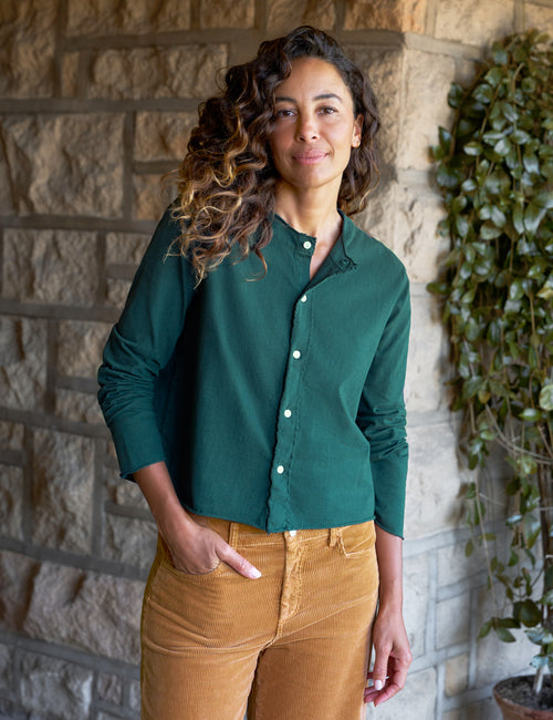 A woman wearing a dark green long-sleeve button-up shirt and tan corduroy pants, standing in front of a stone wall with a green plant to the side., front view