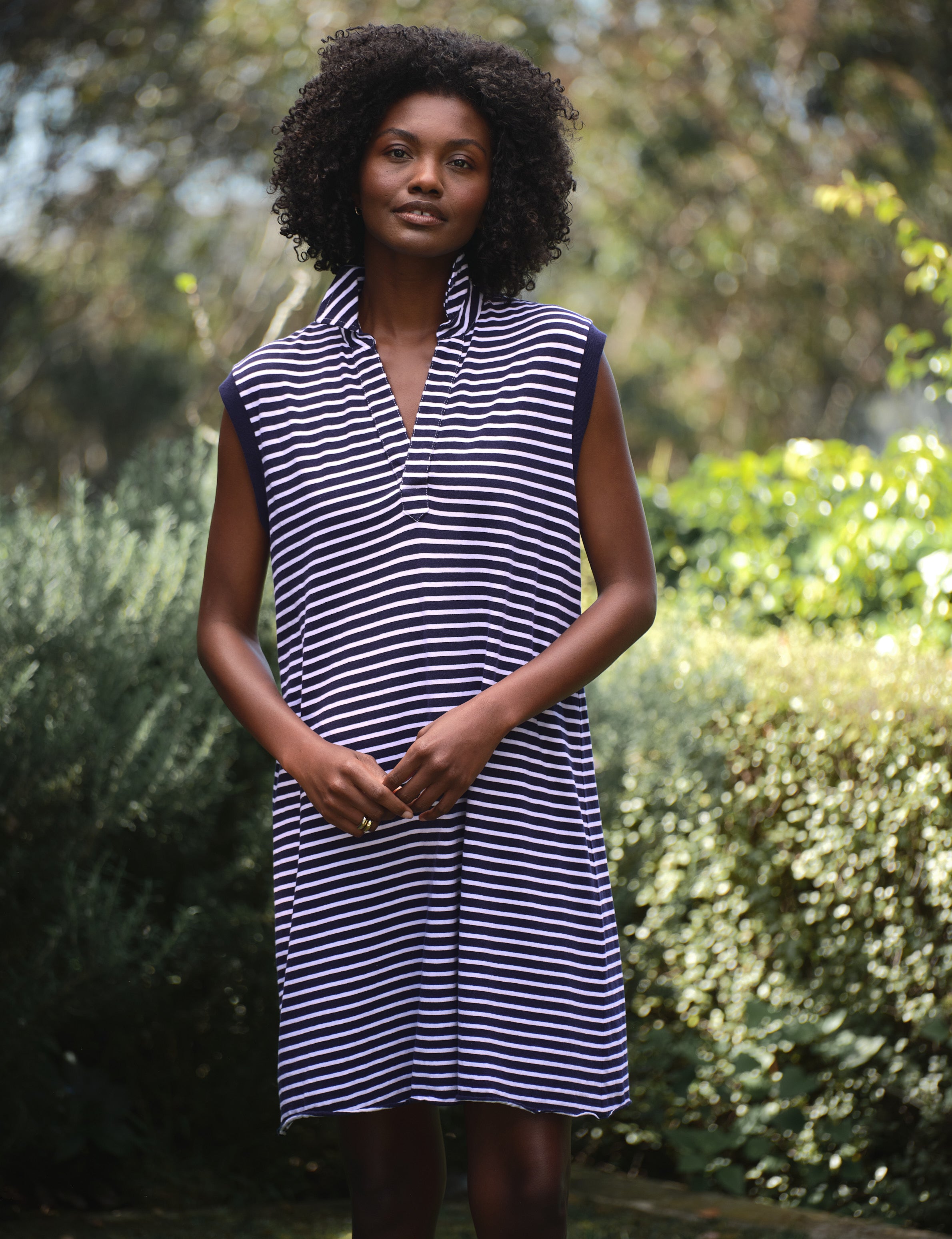 Woman wearing a navy and white striped dress standing outdoors with greenery in the background, view 5
