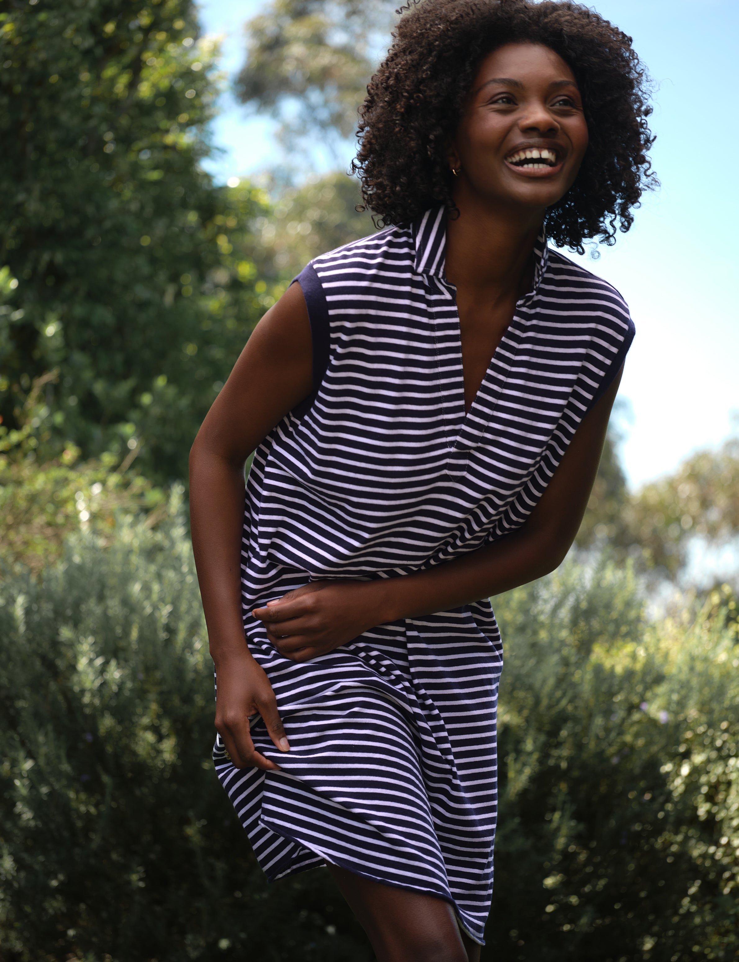 Woman wearing a navy and white striped dress standing outdoors with greenery in the background, view 3