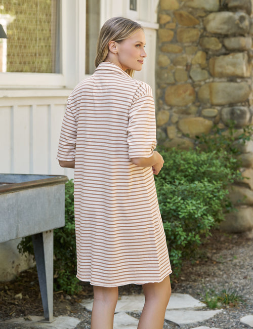A woman wearing a beige and white striped short-sleeve shirt dress, photographed from the back in an outdoor setting with a stone wall and greenery in the background., alternate view