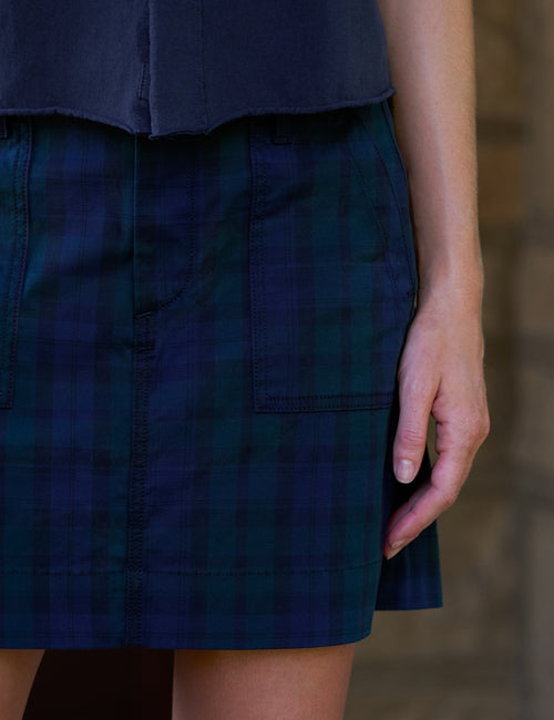 Close-up of a person wearing a dark blue and green plaid mini skirt with a navy top, standing against a blurred stone wall., view 3