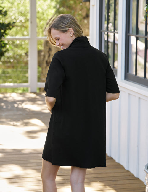 Woman wearing a black dress on a wooden deck with greenery in the background, alternate view