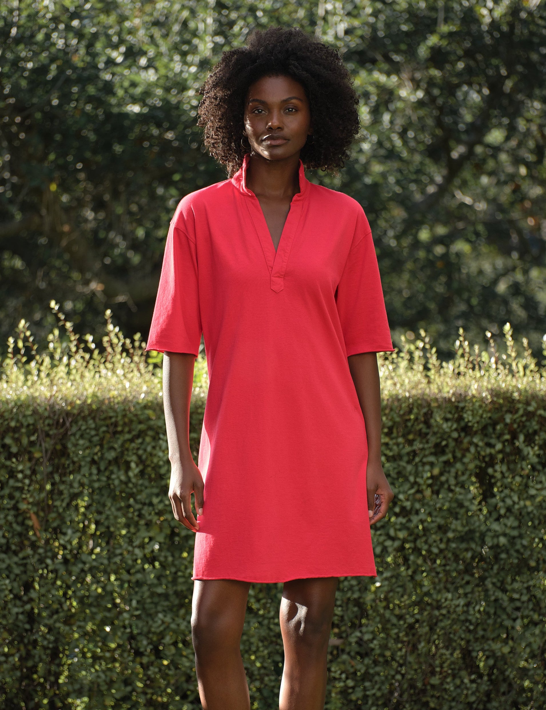 Woman wearing a red dress standing outdoors with greenery in the background, front view