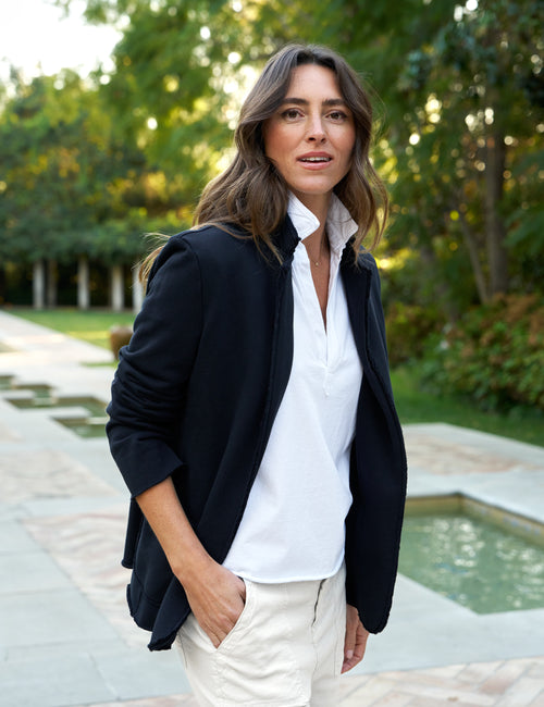 A woman wearing a navy blazer with a white shirt and beige pants standing near a water feature and greenery., front view