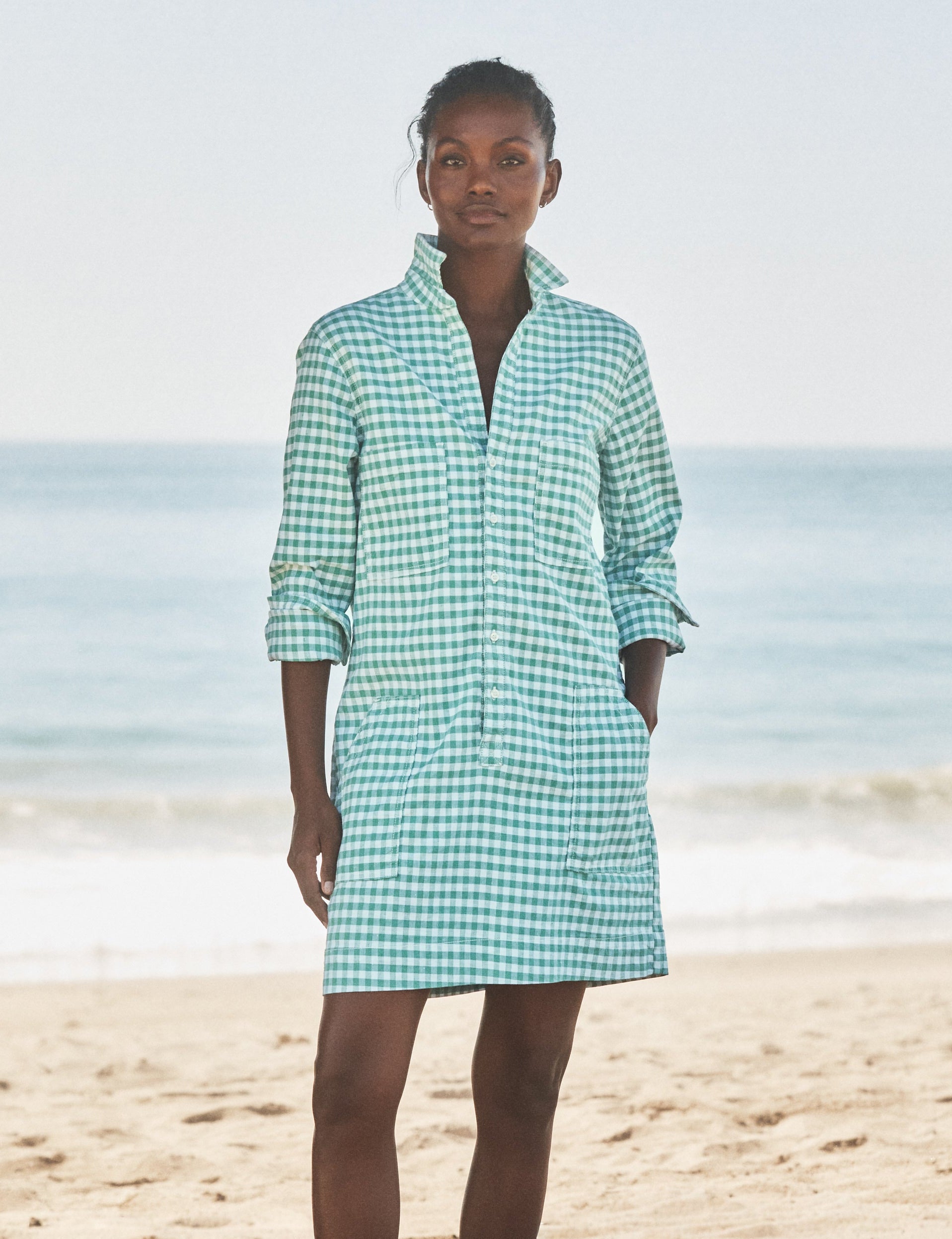Woman wearing a green checkered dress standing on a beach with ocean in the background, front view