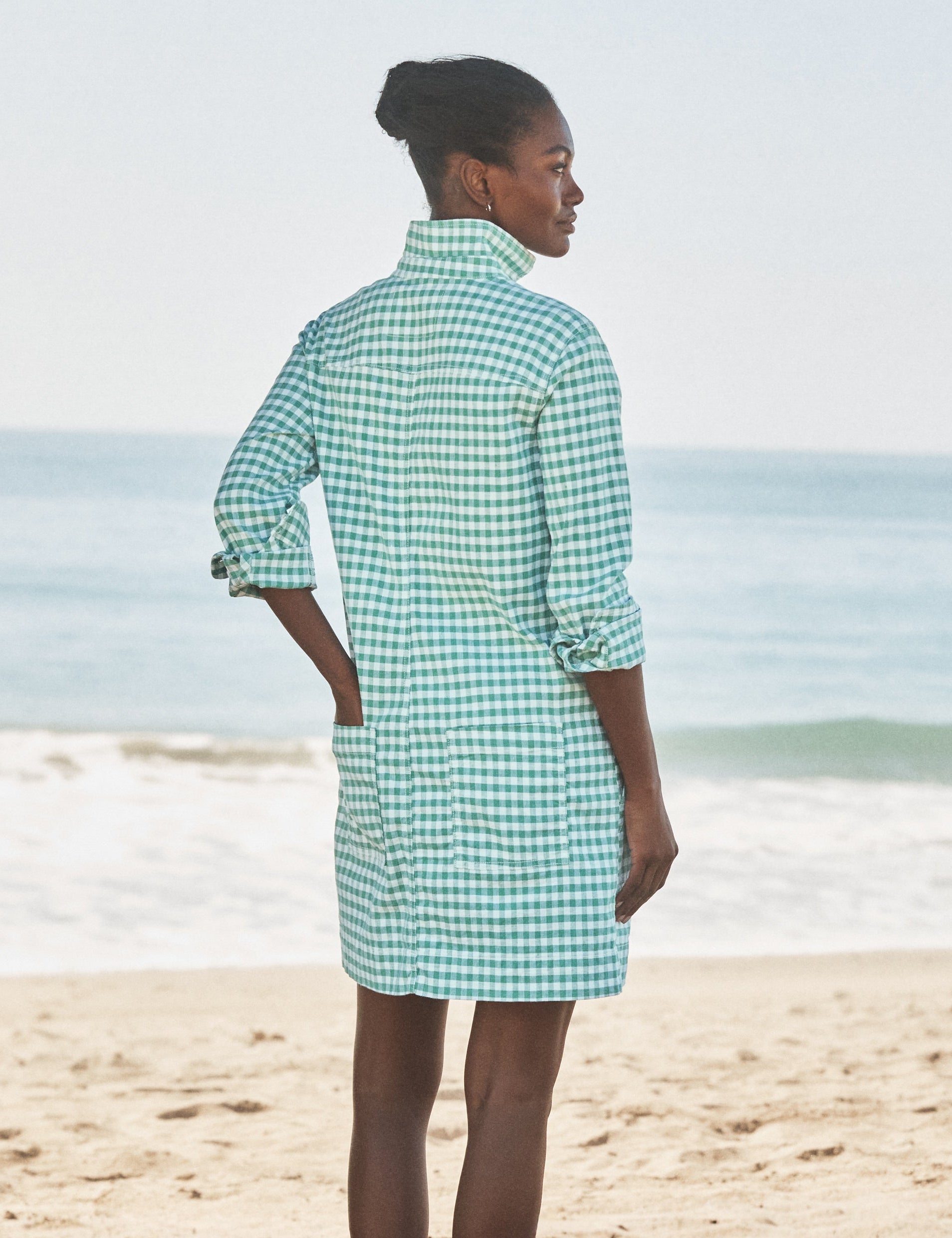 Woman wearing a green checkered dress standing on a beach., alternate view