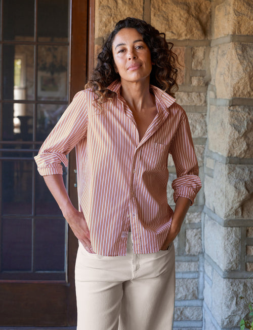 A woman wearing a orange and white vertically striped button-down shirt with beige pants, standing in front of a stone wall and a dark wooden door., front view