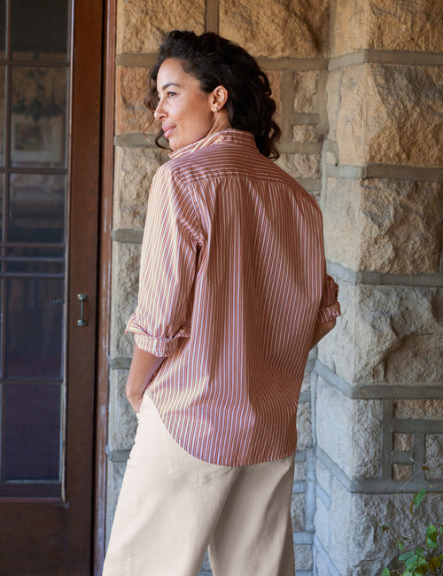 A woman wearing a orange and white vertically striped button-down shirt with rolled-up sleeves and beige pants, standing in front of a stone wall next to a wooden door., alternate view