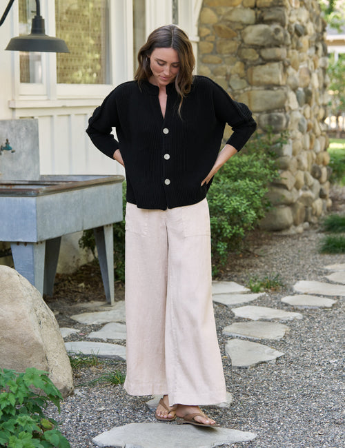 Woman wearing a black cardigan and beige pants standing outdoors near a stone building., view 3