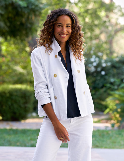 Woman wearing a white jacket with brown buttons and a navy shirt, standing outdoors with greenery in the background., front view