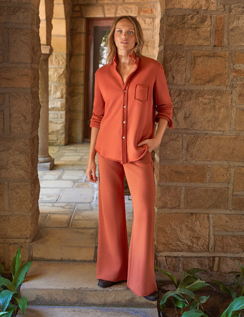 A woman modeling a matching terracotta rust two-piece lounge set consisting of a long-sleeve button-up shirt and wide-leg pants, standing in a stone corridor., front view