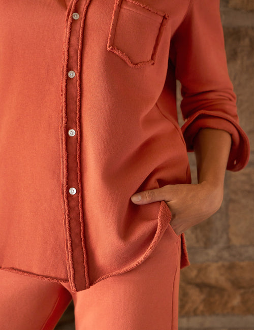 A close-up of a person wearing a rust-orange long-sleeve button-front top with a chest pocket and matching pants, standing against a blurred stone wall., view 3