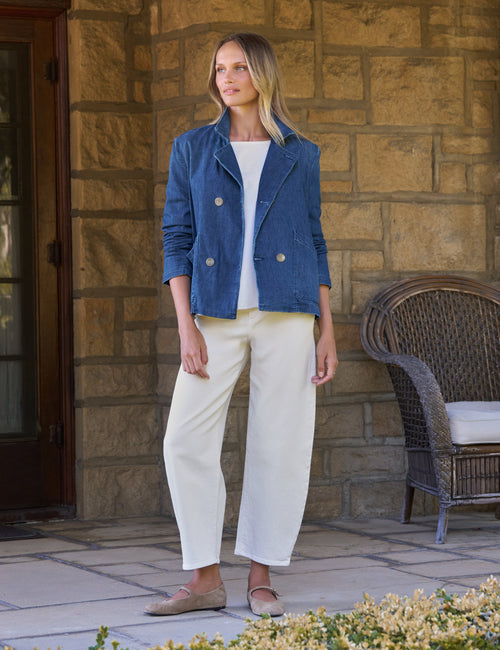 A woman stands on a stone patio wearing a blue double-breasted blazer over a white top, cream cropped trousers, and tan flats. A wicker chair and tan stone wall are in the background., view 5