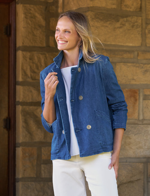 A woman wearing a blue denim jacket over a white top and white pants, standing in front of a tan stone wall., front view