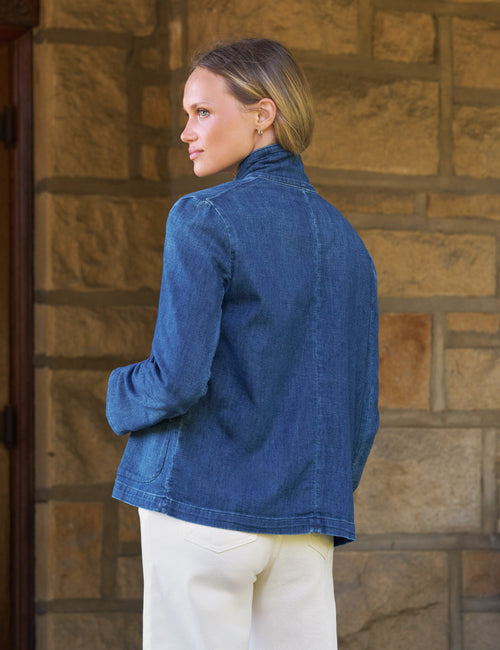 A woman wearing a blue denim jacket and cream pants, shown from the back and side profile against a tan stone wall., alternate view